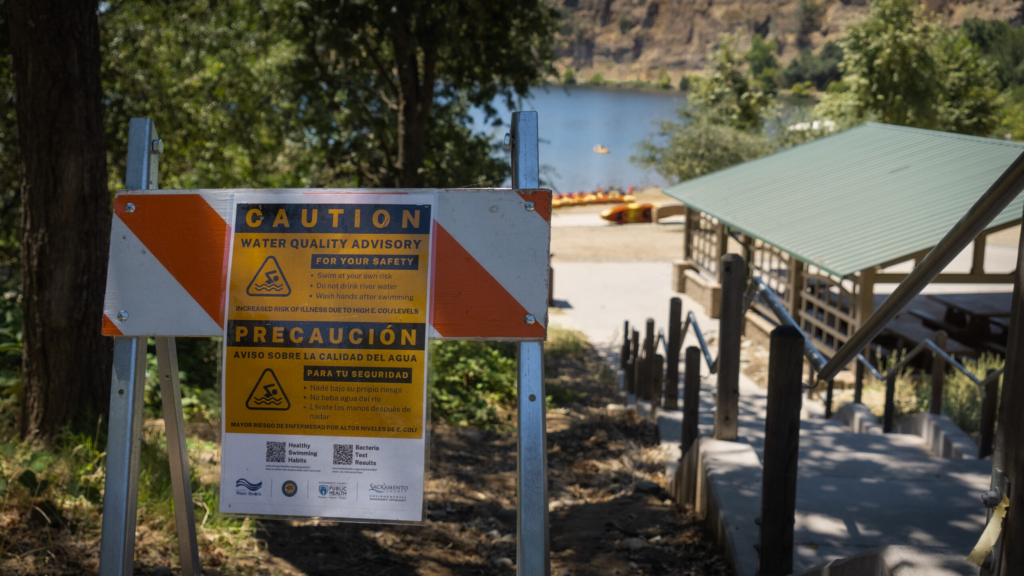 A caution sign with a water quality advisory warning is positioned at the top of one of the entrances to the Black Miners Bar Swim Area at Lake Natoma on July 17, 2026. Virginia Michel